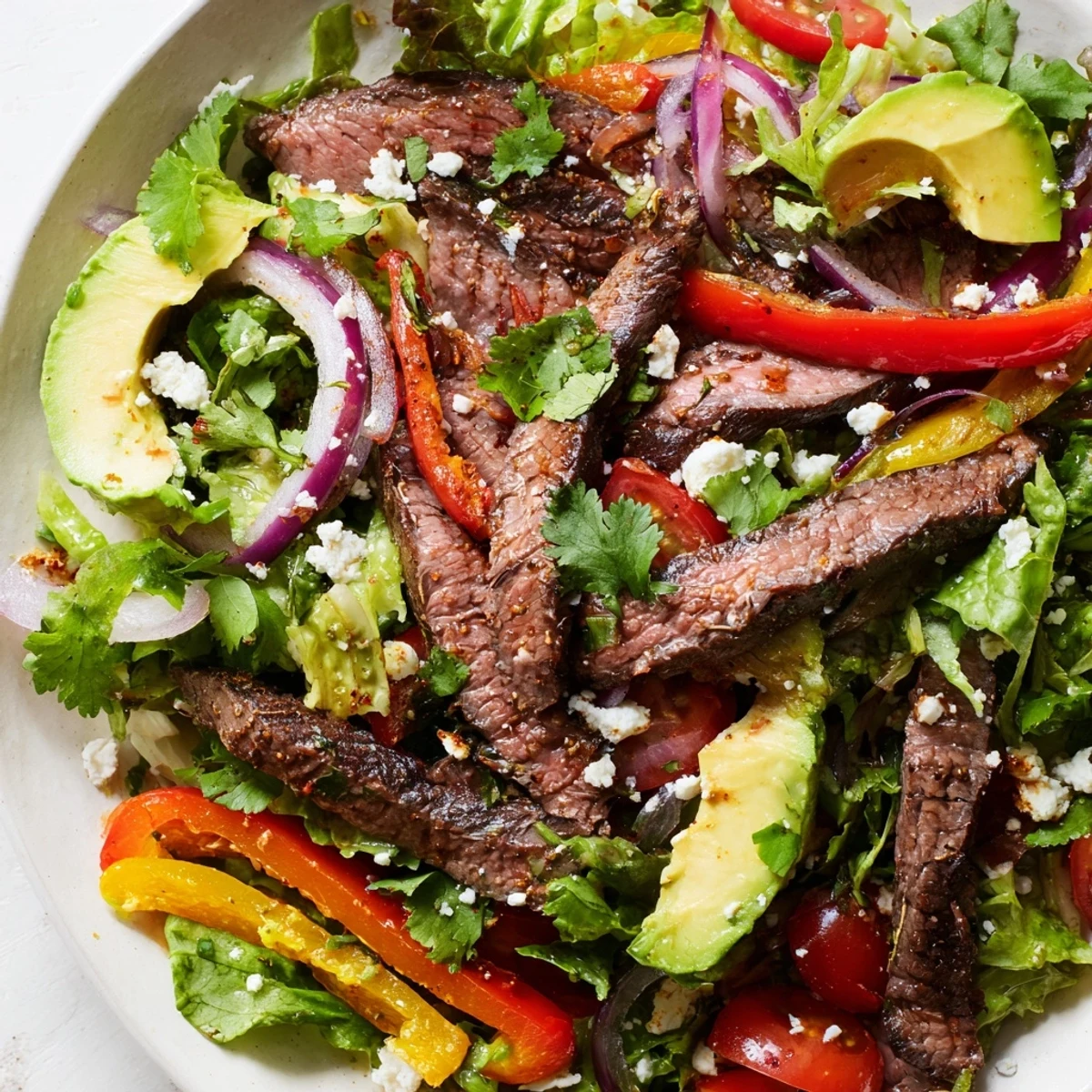 Sliced avocado, cherry tomatoes, and crumbled feta cheese garnish a bowl of Beef Fajita Salad, ready for a generous drizzle of zesty lime vinaigrette.