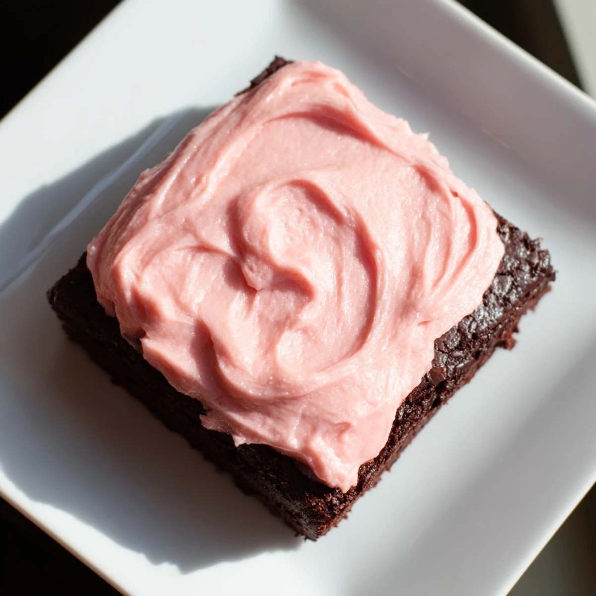 A close-up of frosted brownies shows smooth pink icing topping rich chocolate squares on a cooling rack.  