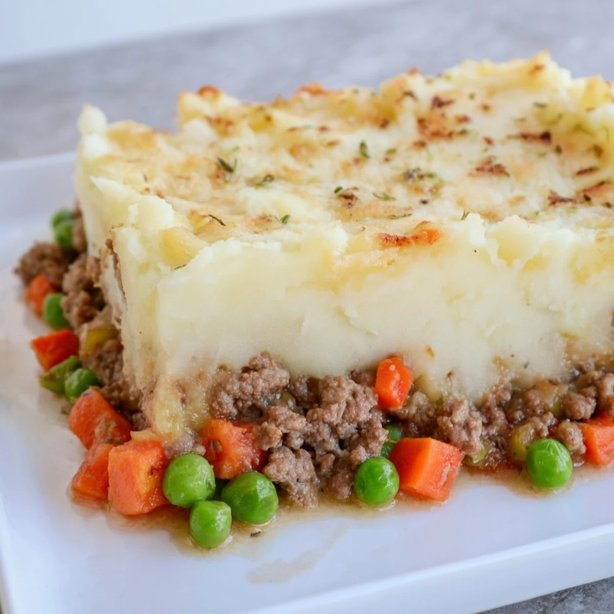 A close-up of Beef Shepherds Pie with Peas reveals golden mashed potato peaks over a bubbling beef and pea filling in a white dish.