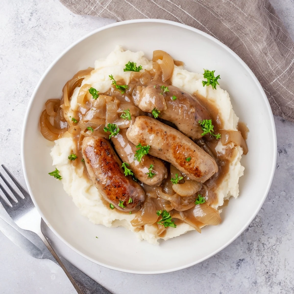 Golden brown sausages nestled in thick onion gravy alongside fluffy mashed potatoes on a plate.