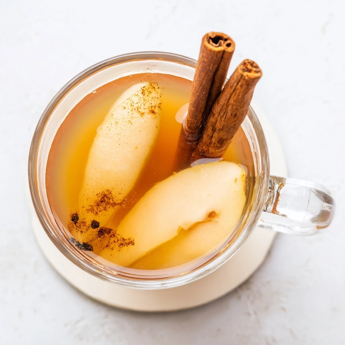 Warm spiced cinnamon pear cider poured into clear glass mugs surrounded by fresh sliced pears and whole cinnamon spices