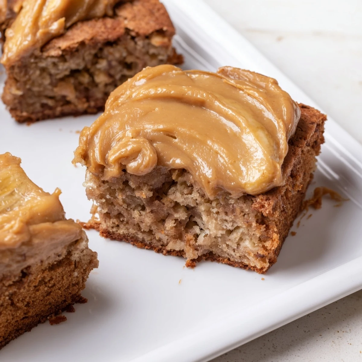 Square slice of banana bread brownies topped with caramelized brown butter frosting on a white dessert plate
