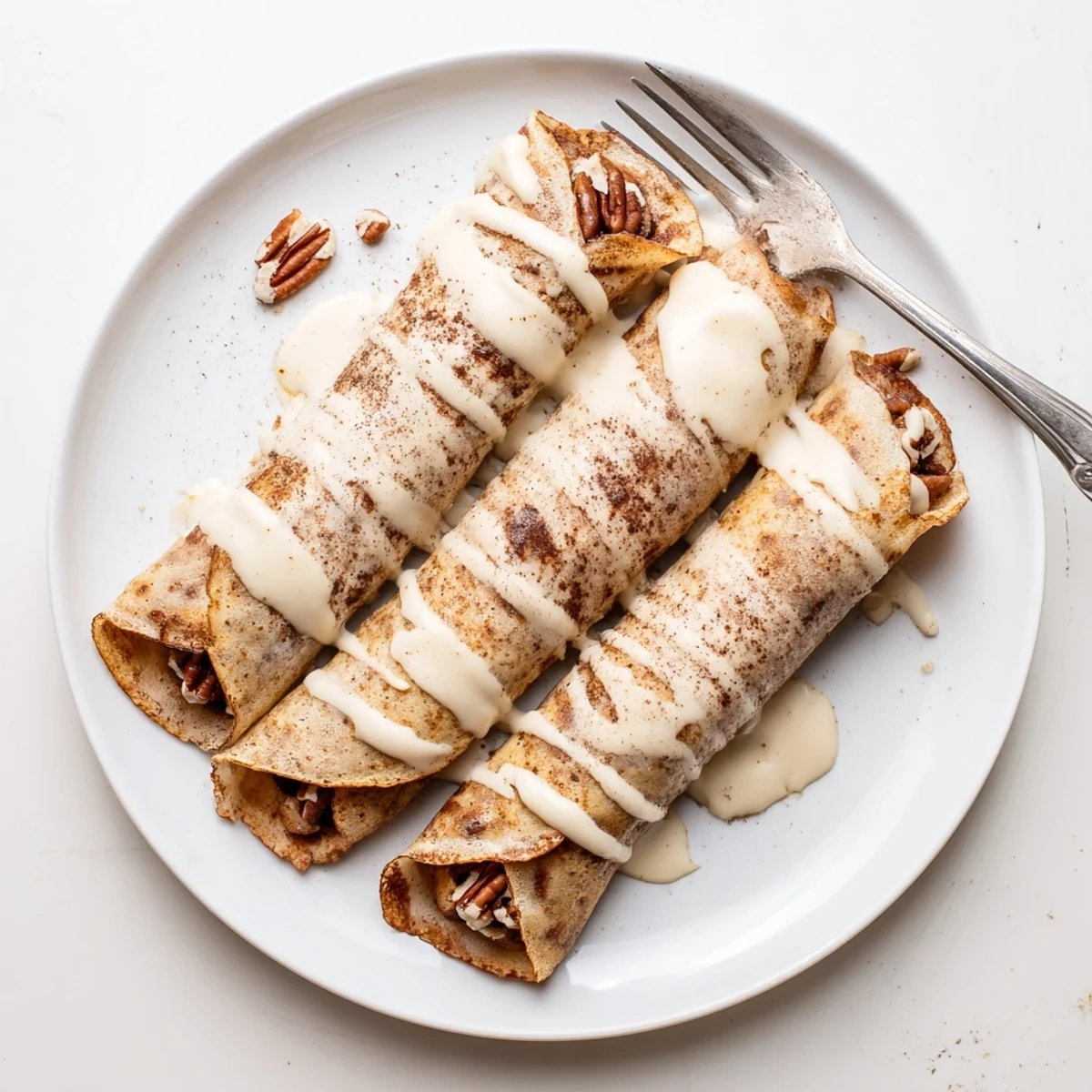 Thin cinnamon roll protein crepes rolled tight and drizzled with cream cheese glaze on a plate